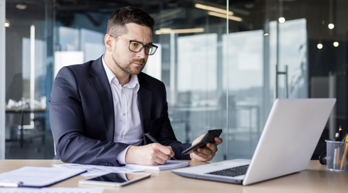 Serious young man accountant, financier and economist working in office at laptop and with documents, holding calculator in hands, concentrating on accounts.