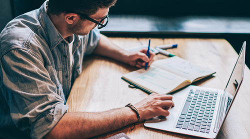 Casual geklede hipster guy schrijven organisatieplan in leerboek voor het onderwijs zittend op desktop met netbook, jonge man schrijven essay in notebook en voorbereiding voor academische test met moderne technologie